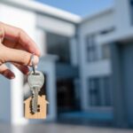 Hand holding a metal key with a small wooden house keychain in front of a modern white house in the background.