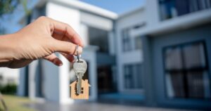 Hand holding a metal key with a small wooden house keychain in front of a modern white house in the background.