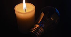 Lit ivory candle beside a tilted incandescent bulb against a dark background, warm and moody.