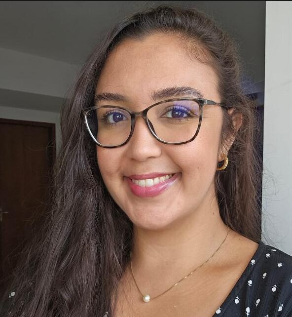 Portrait of a young woman with long dark hair wearing glasses, smiling at the camera indoors, with a pearl necklace and small hoop earring in a casual outfit.