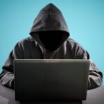 A hooded figure with face hidden, typing at a laptop against a teal background as a cybersecurity/hacking motif.