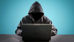 A hooded figure with face hidden, typing at a laptop against a teal background as a cybersecurity/hacking motif.