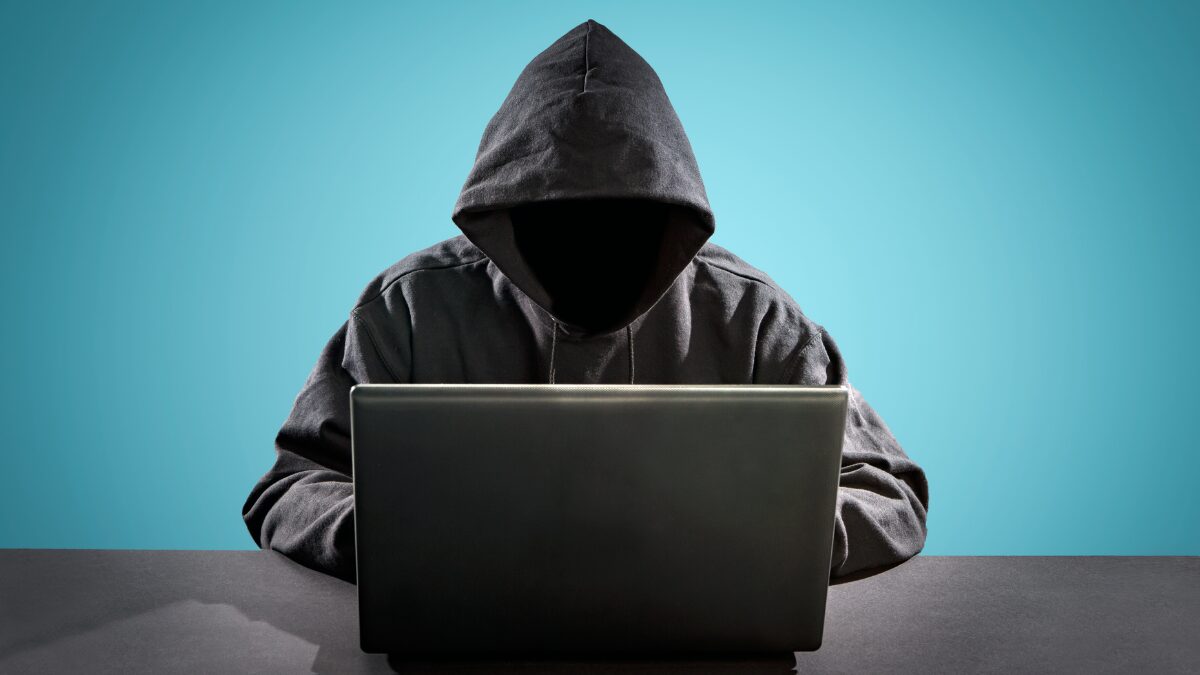 A hooded figure with face hidden, typing at a laptop against a teal background as a cybersecurity/hacking motif.