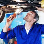 Mechanic in blue overalls inspecting the undercarriage of a raised car with a work light flashlight above him in a shop.