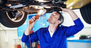 Mechanic in blue overalls inspecting the undercarriage of a raised car with a work light flashlight above him in a shop.