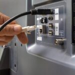 Hand plugging an Ethernet cable into the back panel of a monitor/TV with HDMI and USB ports visible (network connection setup)