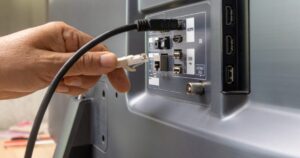 Hand plugging an Ethernet cable into the back panel of a monitor/TV with HDMI and USB ports visible (network connection setup)